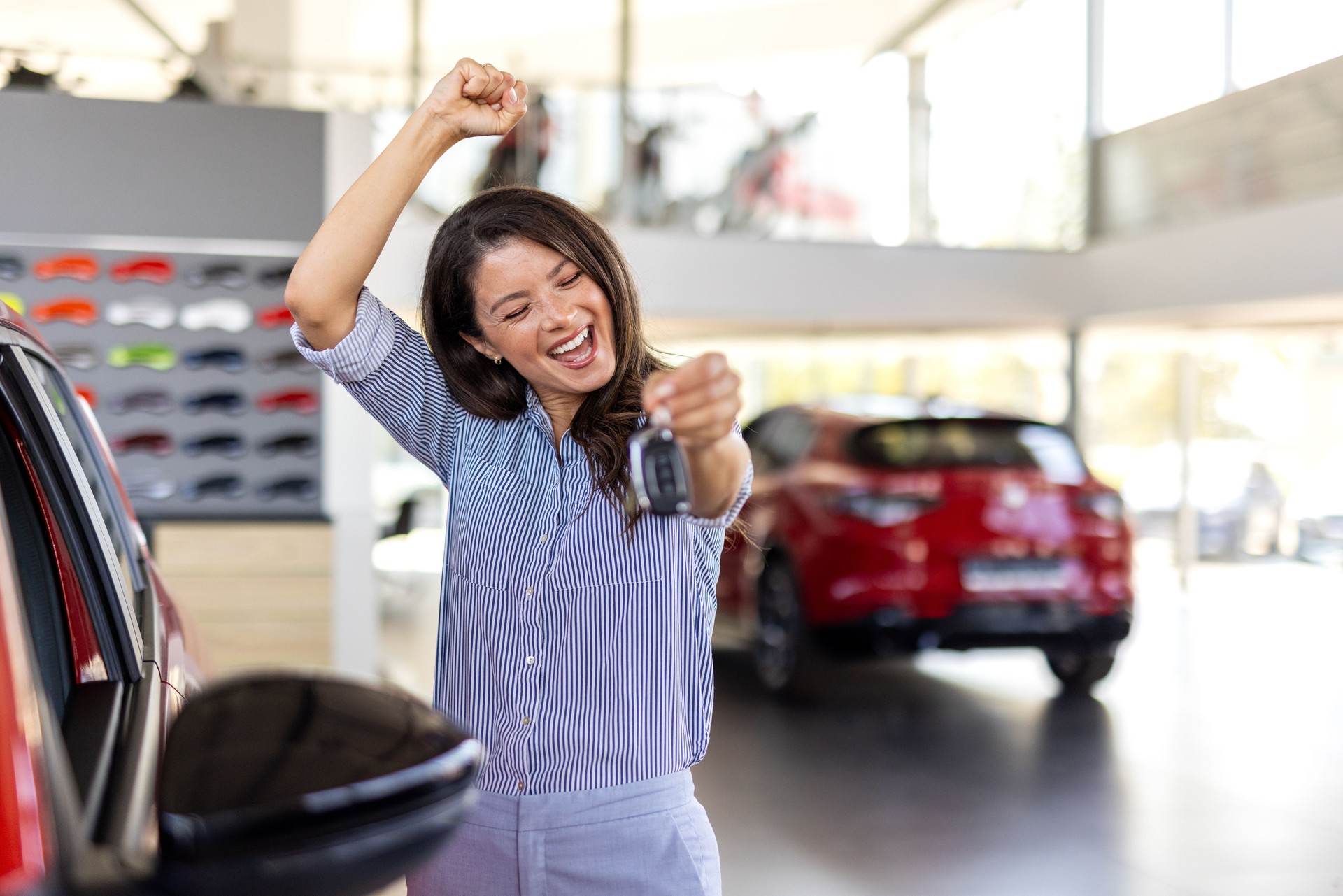 Positive female in smart casual clothes smiling for camera and demonstrating keys while standing near new vehicle in showroom